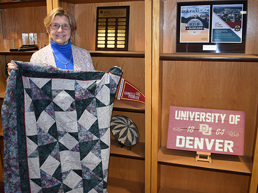Carol Johnson holding a quilt in front of a bookshelf with University of Denver sign