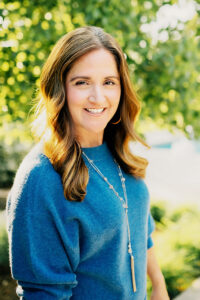 Headshot of Beth Flambures in a blue sweater with trees in the background