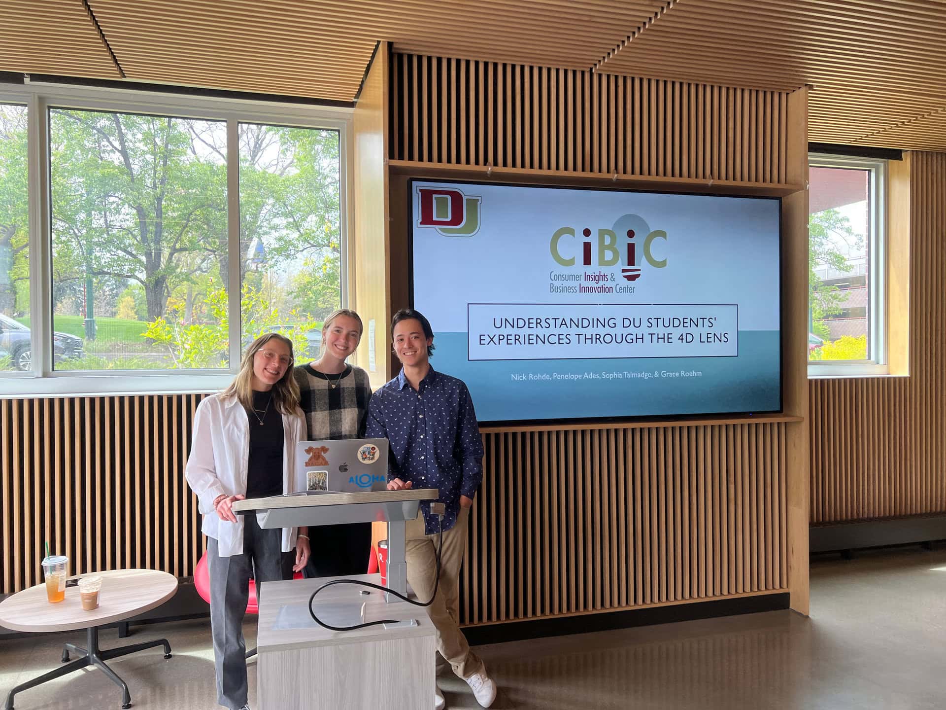 Students posing behind a laptop and monitor displaying a presentation title slide: "Understanding DU Students' Experiences Through the 4D Lens"