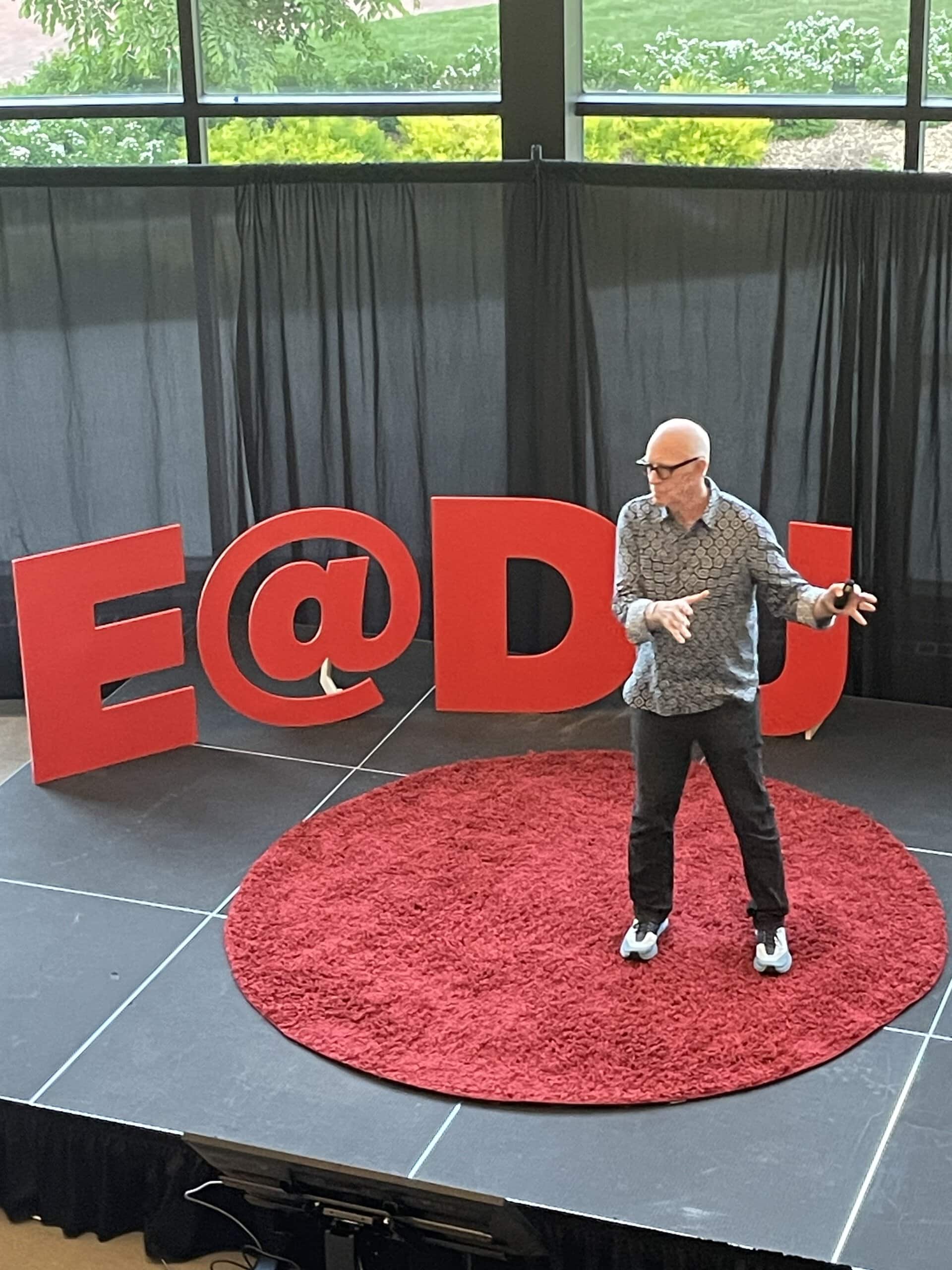 A speaker gesturing on a round red carpet in front of cut out E@DU letters, emulating a TedX talk.