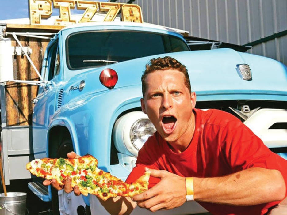 Alumnus Ryan McLean poses in front of his food truck with a giant slice of pizza.