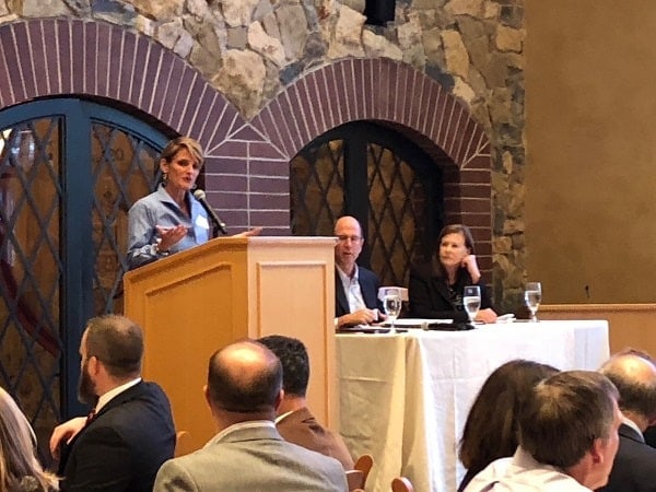 Panel discussion at a Daniels College of Business finance forum, with a female speaker at a podium addressing attendees seated at tables in a stone-arched event venue.